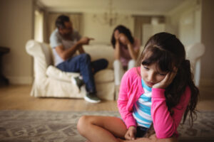 Image of child sitting on the floor listening to parents argue in the background.